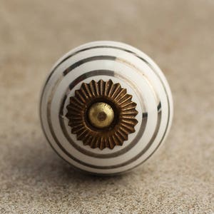 May include: White ceramic knob with silver stripes and a gold metal centre. The knob is round and has a raised design.