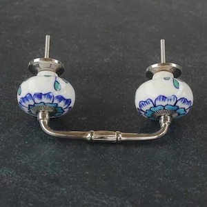 Puede incluir: Un tirador decorativo para armario con dos pomos de cerámica blanca con diseños florales azules. Los pomos están conectados por un tirador de metal plateado. Herrajes diseñados para una fácil instalación.