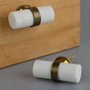 May include: Two white marble drawer pulls with brass accents. The pulls are cylindrical in shape and have a smooth, polished finish.