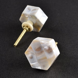 Puede incluir: Dos pomos hexagonales blancos para armario con tornillo dorado. Los pomos tienen un acabado nacarado.