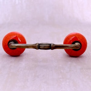 Può includere: Una maniglia per mobili con due sfere in ceramica arancione brillante e strutturata collegate da una barra di metallo color bronzo. Le sfere hanno un effetto craquelé. L'hardware è progettato per mobili.