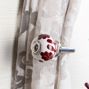 以下が含まれることがあります： 白地に茶色の花柄が描かれたセラミック製のカーテン留め。シルバーの金属製のフックが付いています。