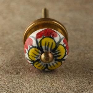 May include: A ceramic knob with a brass base and a floral design. The knob features a yellow flower with red accents on a white background.