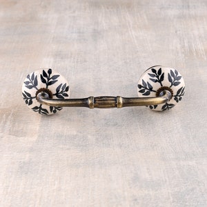 May include: A brass cabinet pull with two white ceramic knobs. The knobs have a black floral design.