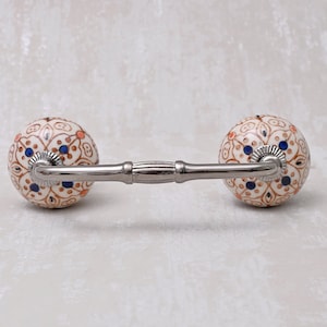 May include: A decorative cabinet knob with a white ceramic base and intricate brown and blue floral patterns. The knob is attached to a silver metal bar.