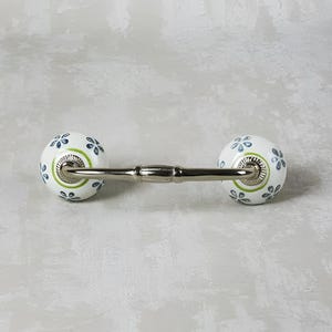 May include: White ceramic drawer pull with a green and blue floral design. The pull has a metal bar connecting the two knobs.