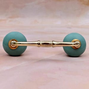 Può includere: Una maniglia per cassetto con due pomelli sferici color verde acqua opaco collegati da una barra in metallo dorato lucido. La barra presenta una texture segmentata simile al bambù e ogni sfera è fissata con una piccola base in metallo dorato rigato.
