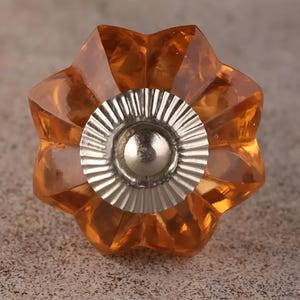 Może przedstawiać: Gwiazdkowaty gałka meblowa z bursztynowego szkła z centralnym elementem ze srebrnego metalu.