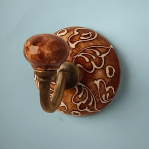 Puede incluir: Un gancho de cerámica marrón con un diseño floral blanco. El gancho tiene un gancho de latón y está montado en una pared.