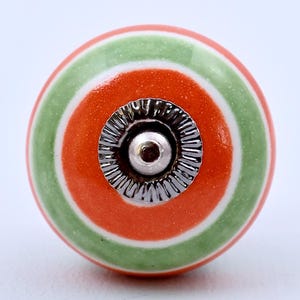 Puede incluir: Un pomo de cerámica redondo para muebles con anillos concéntricos de color naranja brillante y verde salvia suave. El centro del pomo está adornado con una pieza circular de metal plateado que tiene un borde estriado y un centro liso.