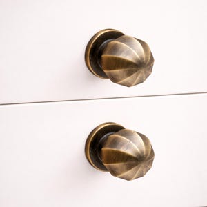 May include: Close-up of two antique brass cabinet knobs with a spiral design. The knobs are mounted on a white surface, showcasing their aged, metallic finish. The knobs have a round base and a textured, faceted center.