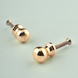Puede incluir: Dos pomos de armario de metal de color dorado con un diseño redondo ligeramente texturizado. Cada pomo tiene un tornillo para sujetarlo a una puerta de armario.