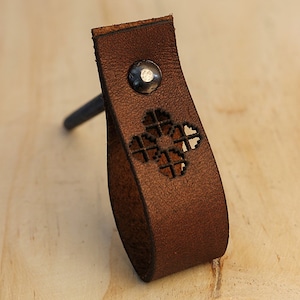 Pode incluir: Uma puxador de armário de couro marrom com um design de corte decorativo e um rebite de metal.
