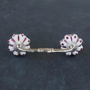 May include: White ceramic drawer pull with a silver metal bar connecting two red and white polka dot flower-shaped knobs.
