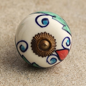 Puede incluir: Pomo de cerámica blanco con un diseño floral verde, azul y rojo. El pomo tiene un tornillo de metal dorado en el centro.