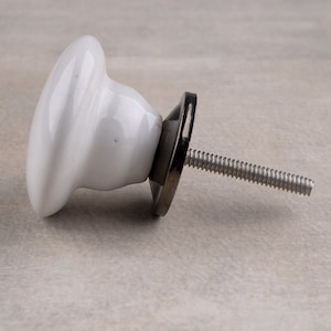 May include: A white ceramic cabinet knob with a smooth, rounded mushroom-shaped head. The knob features a dark metallic base plate and a threaded metal bolt extending from the back, designed for attachment to furniture or cabinetry doors.