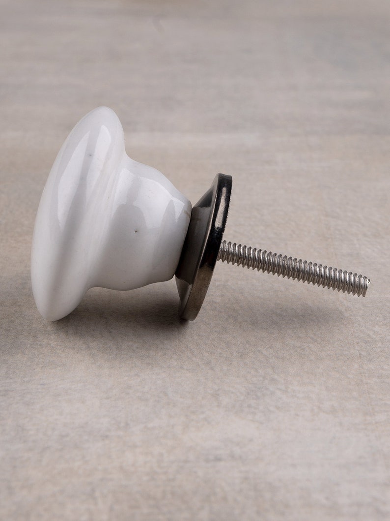 May include: A white ceramic cabinet knob with a smooth, rounded mushroom-shaped head. The knob features a dark metallic base plate and a threaded metal bolt extending from the back, designed for attachment to furniture or cabinetry doors.
