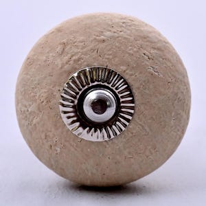 Peut inclure: Un bouton de meuble rond de couleur beige avec une surface texturée rappelant la pierre. Le centre présente une plaque métallique circulaire argentée avec un motif strié en forme de soleil autour d'un petit bouton métallique lisse.