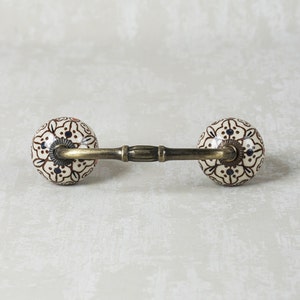 May include: A decorative cabinet pull with a brown and white floral design. The pull has a brass bar connecting two ceramic knobs.
