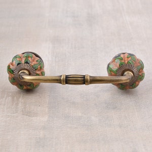 Könnte beinhalten: Ein dekorativer Schrankgriff mit Messing-Finish und zwei Keramikknöpfen. Die Knöpfe sind orange, grün und schwarz mit einem floralen Muster.