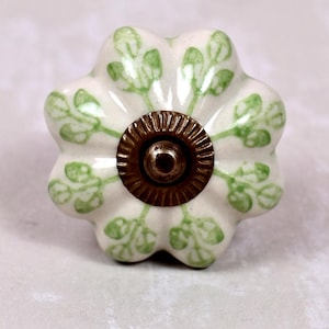 May include: A flower-shaped ceramic cabinet knob with a cream-colored base and a hand-painted green leaf pattern. The center features a circular, ribbed, antique bronze-toned metal hardware piece with a small central screw head for mounting.