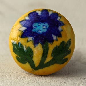 May include: A yellow ceramic knob featuring a blue and green floral design. The flower has a white centre and blue petals.