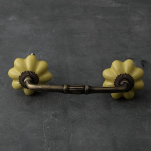 May include: A yellow ceramic drawer pull with a brass arch-shaped handle. The pull has a flower-like design.
