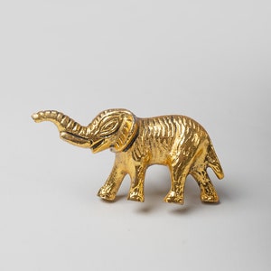May include: A gold-colored metal elephant figurine with a raised trunk. The elephant is standing on a white surface.
