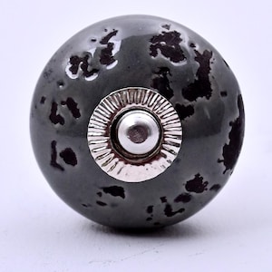 May include: A round cabinet knob with a glossy dark gray finish featuring irregular black mottled spots. The center of the knob has a circular silver-toned metal plate with a ridged edge and a smooth, raised silver-toned metal dome in the middle.