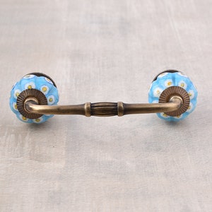 May include: A decorative cabinet pull with blue ceramic knobs featuring a white and yellow floral pattern. The pull has a bronze-colored metal bar and mounting hardware, adding a touch of vintage charm to furniture or cabinetry.