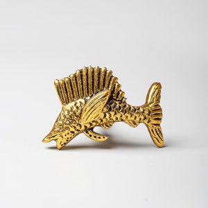 Könnte beinhalten: Eine goldfarbene Metallfigur eines Fisches mit detailliertem Design. Der Fisch hat eine lange, spitze Schnauze und eine Rückenfinne, die wie ein Kamm geformt ist. Der Fisch befindet sich in einer Sprungposition, mit dem Schwanz nach oben.