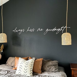 May include: A bedroom with a dark grey accent wall featuring the text "always kiss me goodnight" in white. The room has a large bed with a grey duvet and brown pillows. Two wicker pendant lights hang from the ceiling.