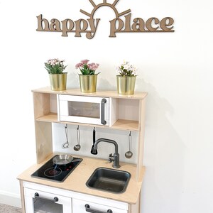 Op de afbeelding: Een houten speelkeuken met een witte kast, een zwart fornuis en een zwarte gootsteen. De keuken heeft een wit rek boven het fornuis met een kleine oven. De keuken is versierd met een houten bord waarop "happy place" staat met een zon boven de woorden.