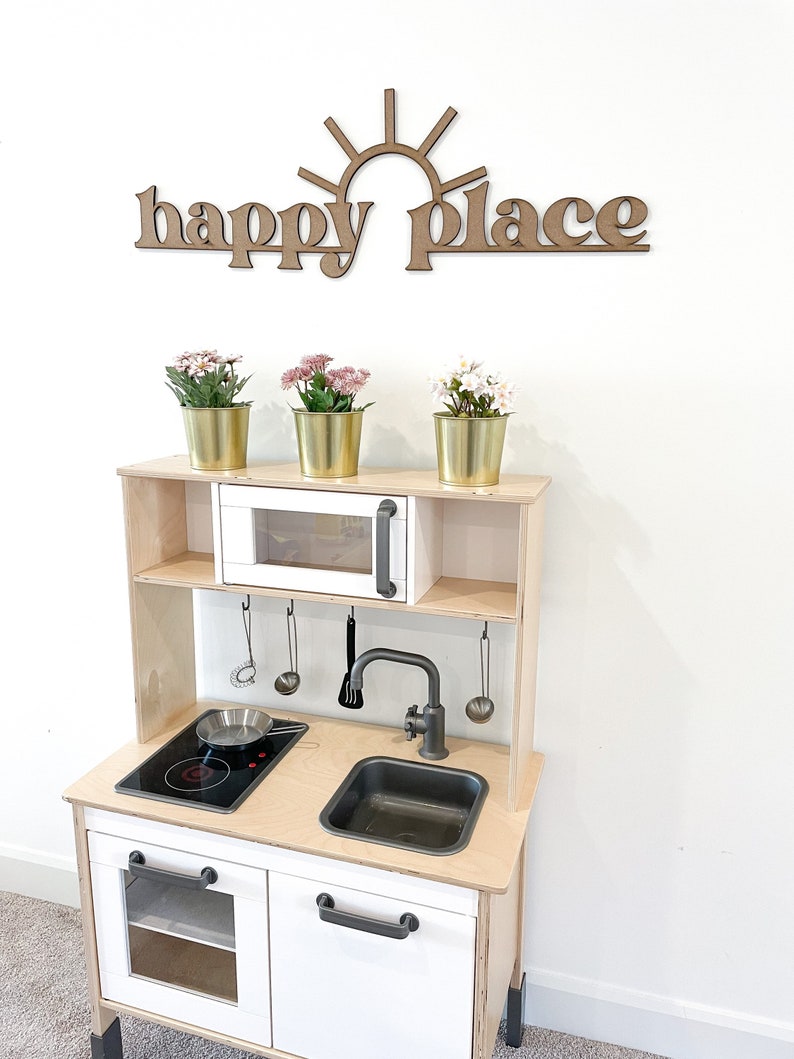 Op de afbeelding: Een houten speelkeuken met een witte kast, een zwart fornuis en een zwarte gootsteen. De keuken heeft een wit rek boven het fornuis met een kleine oven. De keuken is versierd met een houten bord waarop "happy place" staat met een zon boven de woorden.