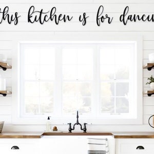 May include: A well-lit kitchen featuring white units, wooden worktops, and open shelving. The phrase "this kitchen is for dancing" is displayed above the window. Decorative items include plants, crockery, and chopping boards, creating a warm and inviting atmosphere.