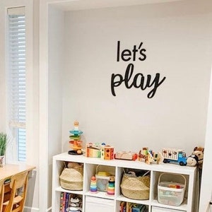 May include: A white wall featuring black script lettering that reads let's play, positioned above a white cubby storage unit filled with various colourful toys, woven baskets, and books, creating an organised space for children's activities.