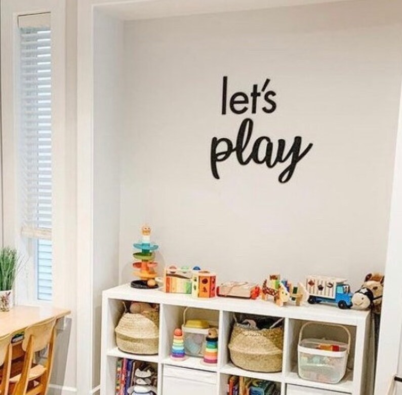 May include: A white wall featuring black script lettering that reads let's play, positioned above a white cubby storage unit filled with various colourful toys, woven baskets, and books, creating an organised space for children's activities.