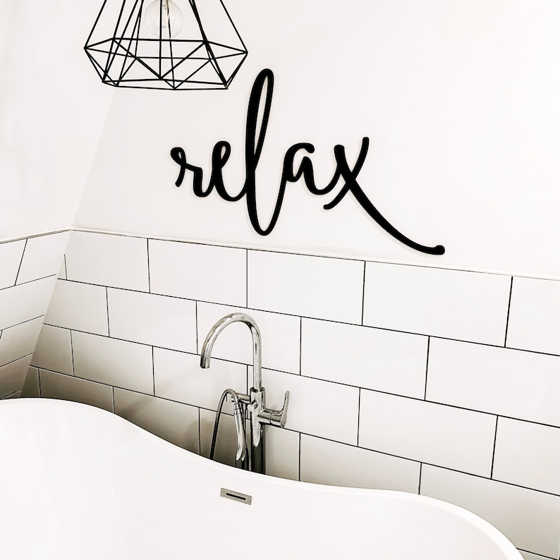 May include: A white bathroom with white subway tile and a freestanding bathtub. A black metal sign with the word "relax" is mounted on the wall above the tub. A chrome faucet is attached to the tub.