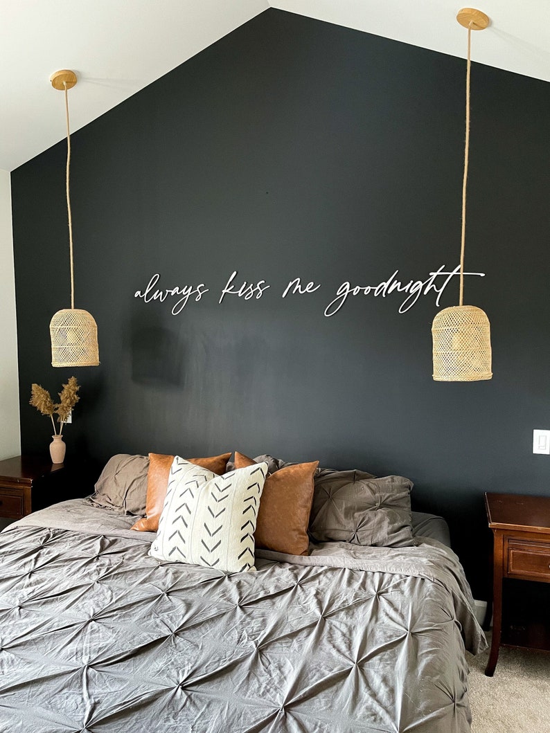 May include: A bedroom with a dark grey accent wall featuring the text "always kiss me goodnight" in white. The room has a large bed with a grey duvet and brown pillows. Two wicker pendant lights hang from the ceiling.
