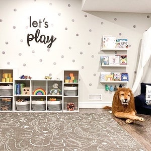 May include: A playroom featuring a white cube storage unit with fabric bins, a grey dinosaur-patterned rug, and wall-mounted shelves holding books. The wall displays silver polka dots and black text reading let's play, with a white canopy and lion plush.