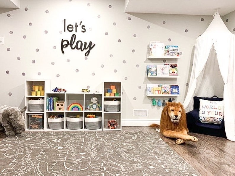 May include: A playroom featuring a white cube storage unit with fabric bins, a grey dinosaur-patterned rug, and wall-mounted shelves holding books. The wall displays silver polka dots and black text reading let's play, with a white canopy and lion plush.