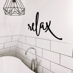May include: A white bathroom with white subway tile and a white bathtub. A black metal sign with the word "relax" is mounted on the wall above the bathtub. A chrome faucet is attached to the bathtub.
