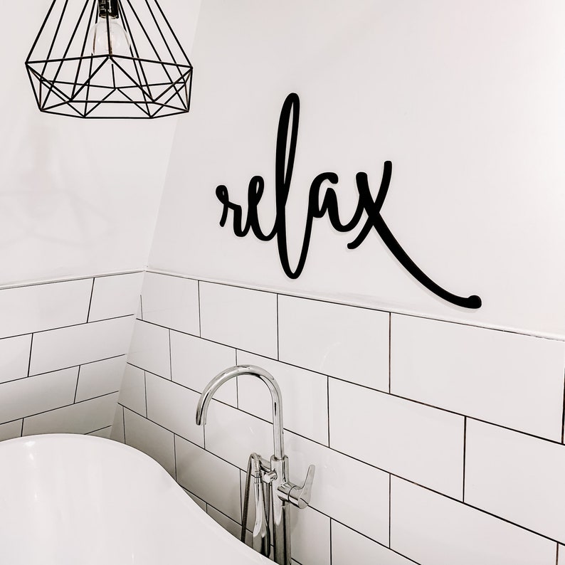 May include: A white bathroom with white subway tile and a white bathtub. A black metal sign with the word "relax" is mounted on the wall above the bathtub. A chrome faucet is attached to the bathtub.