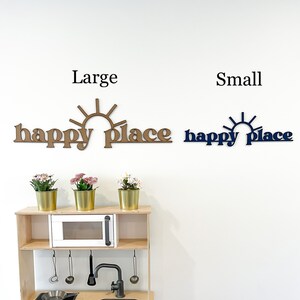 Op de afbeelding: Twee houten wandborden met de tekst "happy place" en een zonafbeelding. Het bord links is groter en bruin, terwijl het bord rechts kleiner en blauw is. De borden hangen aan een witte muur boven een houten speelkeuken.