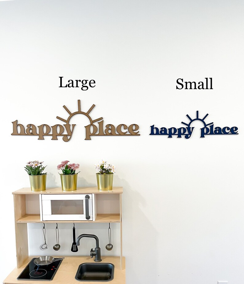 Op de afbeelding: Twee houten wandborden met de tekst "happy place" en een zonafbeelding. Het bord links is groter en bruin, terwijl het bord rechts kleiner en blauw is. De borden hangen aan een witte muur boven een houten speelkeuken.