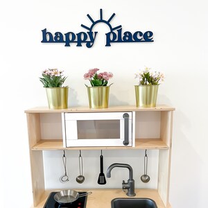 Op de afbeelding: Een witte houten speelkeuken met een zwart fornuis, een zwarte gootsteen en een witte magnetron. Op de bovenste plank staan drie gouden potten met roze bloemen. De woorden "happy place" staan in het blauw op de muur boven de keuken.