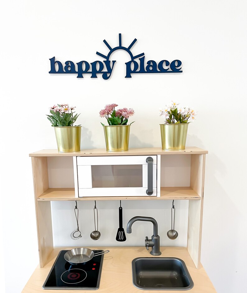 Op de afbeelding: Een witte houten speelkeuken met een zwart fornuis, een zwarte gootsteen en een witte magnetron. Op de bovenste plank staan drie gouden potten met roze bloemen. De woorden "happy place" staan in het blauw op de muur boven de keuken.
