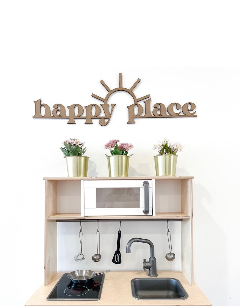Op de afbeelding: Een houten speelkeuken met een witte magnetron, een zwarte kraan en een zwart fornuis. De keuken heeft een plank met drie gouden potten met roze bloemen. Boven de keuken hangt een houten bord met de tekst "happy place" met een zonnetje.