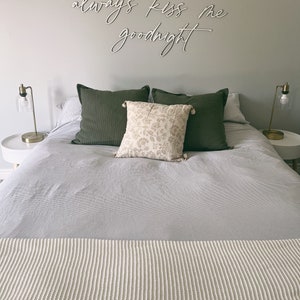 May include: A bedroom with a white bedspread and a white knitted throw blanket. The bed has two green pillows and a floral patterned pillow. The wall above the bed has a white sign that reads "always kiss me goodnight".