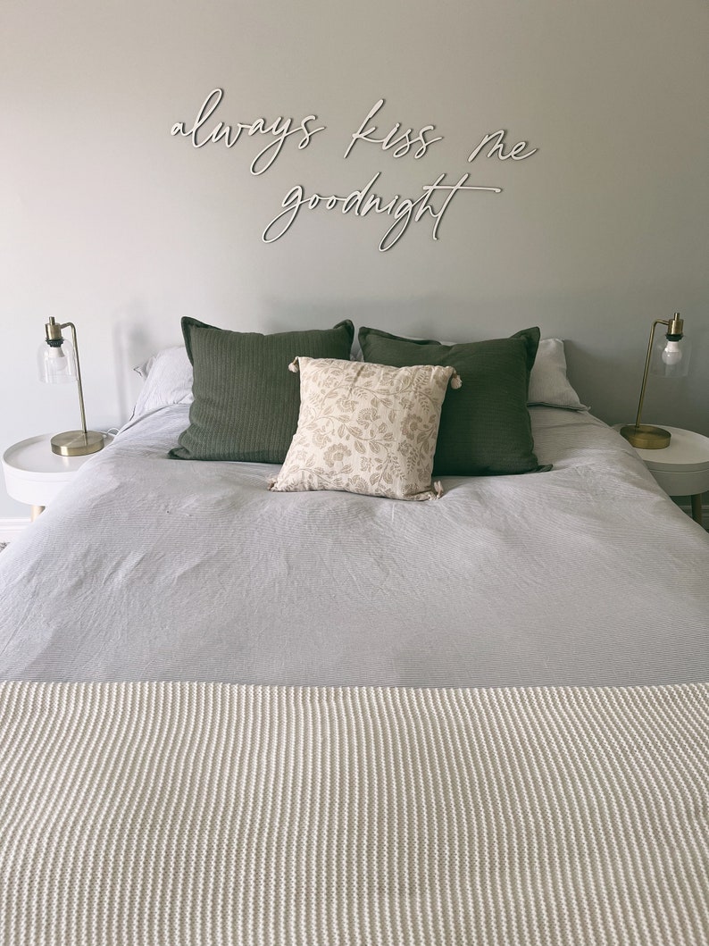 May include: A bedroom with a white bedspread and a white knitted throw blanket. The bed has two green pillows and a floral patterned pillow. The wall above the bed has a white sign that reads "always kiss me goodnight".